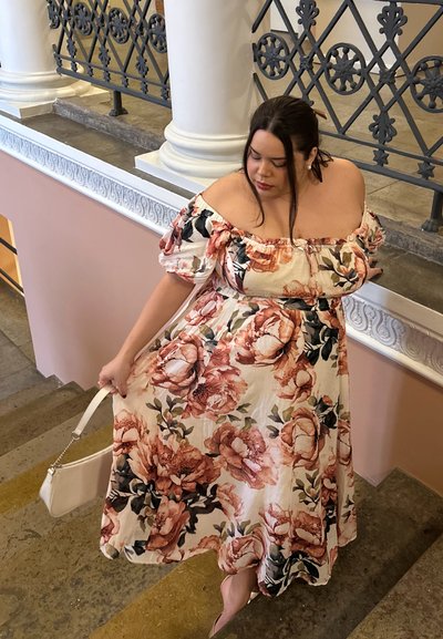 Vestido maxi floral con diseño fuera de los hombros, que presenta grandes flores en tonos durazno y crema. La modelo sostiene un bolso blanco mientras baja por las escaleras.