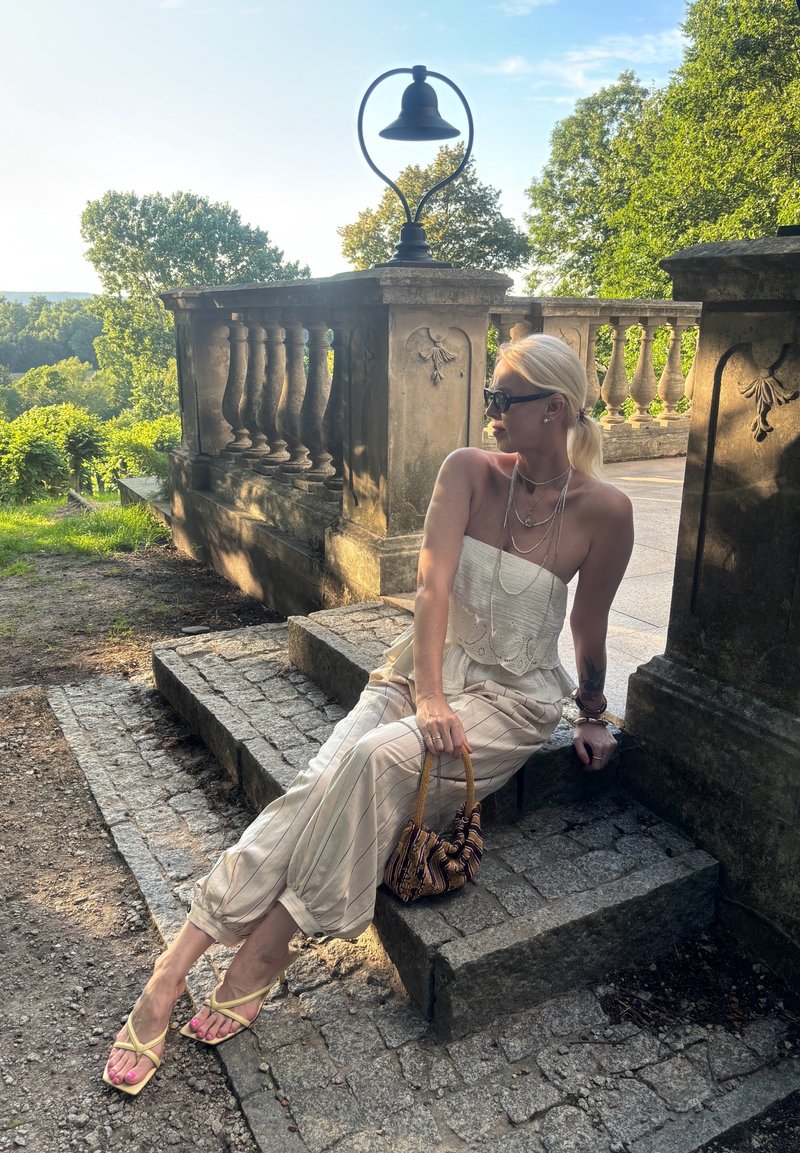 Femme en tenue légère d'été et sandales assise sur des marches en pierre près d'une balustrade ornée dans un jardin extérieur ensoleillé.