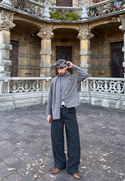 Hombre con suéter gris, bufanda, pantalones a cuadros y gorra, de pie sobre un pavimento de piedra con columnas ornamentadas y barandillas al fondo.