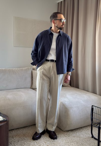 Hombre con camisa azul oscuro, camiseta blanca, pantalones beige y zapatos oscuros, de pie en una sala cerca de un sofá beige y cortinas.