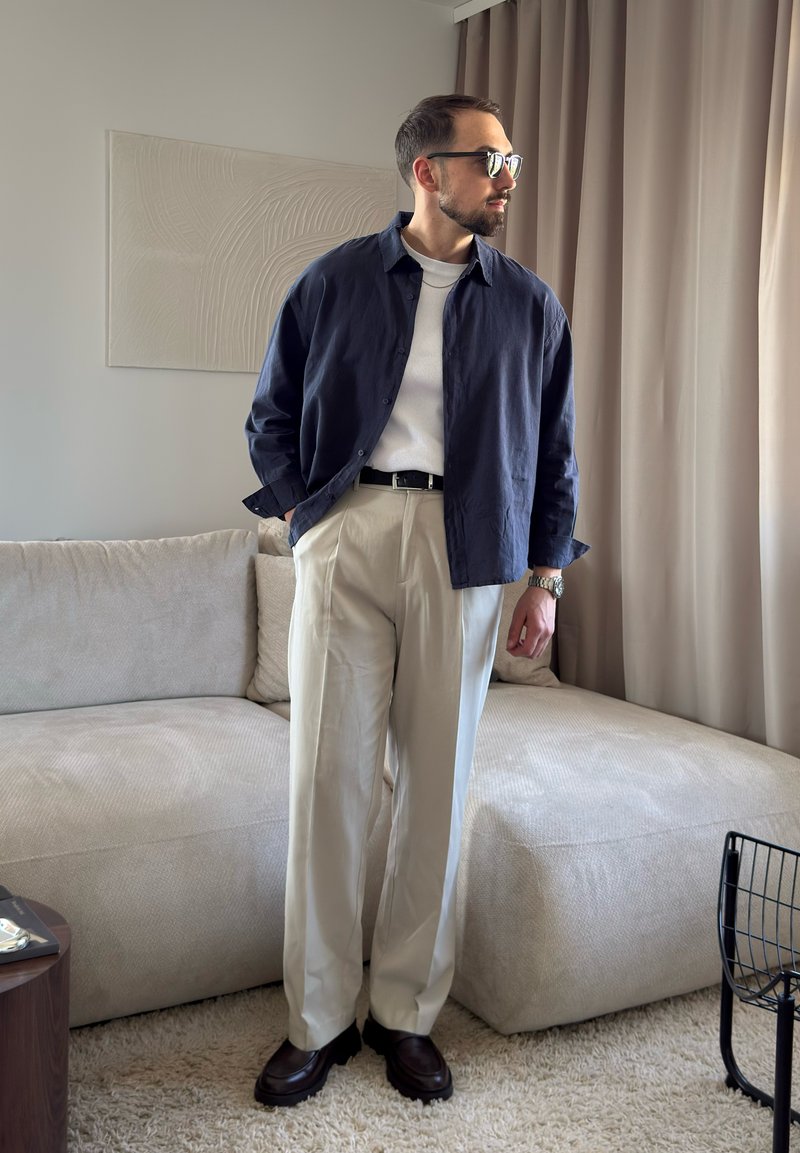 Homme portant une chemise bleu foncé, un t-shirt blanc, un pantalon beige et des chaussures foncées, debout dans un salon près d'un canapé beige et des rideaux.