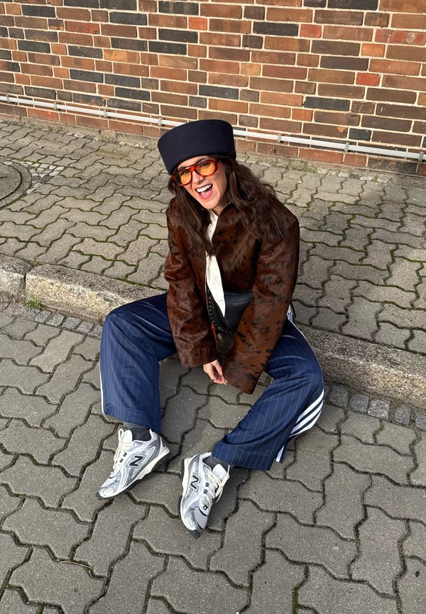 Person som sitter på trottoarkanten, klädd i en mörkbrun prickig jacka, blå randiga byxor, svart hatt, orange solglasögon och vita sneakers.