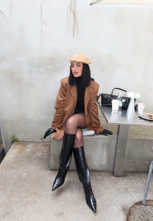 Mujer con boina beige, blazer marrón, guantes negros, botas negras y medias transparentes sentada en un banco de hormigón al aire libre cerca de una mesa de metal con bebidas.