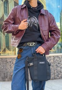 Sac en tissu noir avec une bandoulière réglable, doté d'une boucle, tenu dans une main. Porté avec une veste en cuir bordeaux et un jean en denim.