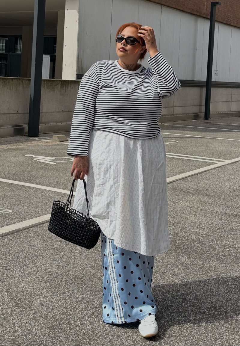 Person mit roten Haaren und Sonnenbrille trägt gestreiftes Oberteil, weißes schürzenähnliches Kleidungsstück, blaue gepunktete Hose und hält eine gewebte schwarze Handtasche auf einem Parkplatz.