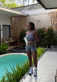 Femme en tenue de sport tenant une bouteille d'eau, debout près d'une piscine avec des plantes et des sièges extérieurs dans une cour ensoleillée.