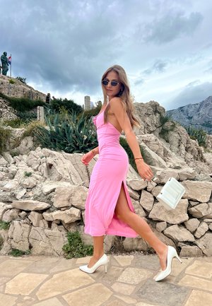Vestido ceñido rosa con abertura lateral, combinado con tacones blancos. La modelo lleva un pequeño bolso blanco y lleva gafas de sol. Fondo rocoso natural.