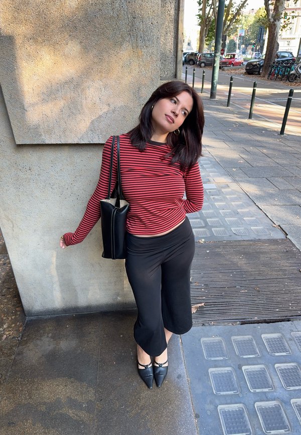 Une femme vêtue d’un haut à rayures rouges et noires et d’un pantalon noir s’appuie contre un mur en pierre sur un trottoir en ville, avec un sac noir en bandoulière.
