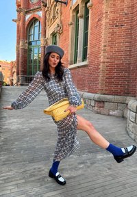 Hosszú, áttetsző, kockás ruha szürkében és fehérben, puffos ujjakkal; egy fonott, sárga clutch-ot tart. Sötét zoknik fekete és fehér cipőkkel. Szürke beret.