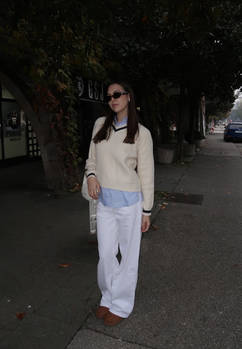 White oversized jumper with navy trim worn over a light blue shirt, paired with white wide-leg trousers and brown slip-on shoes. Black sunglasses and a handbag.
