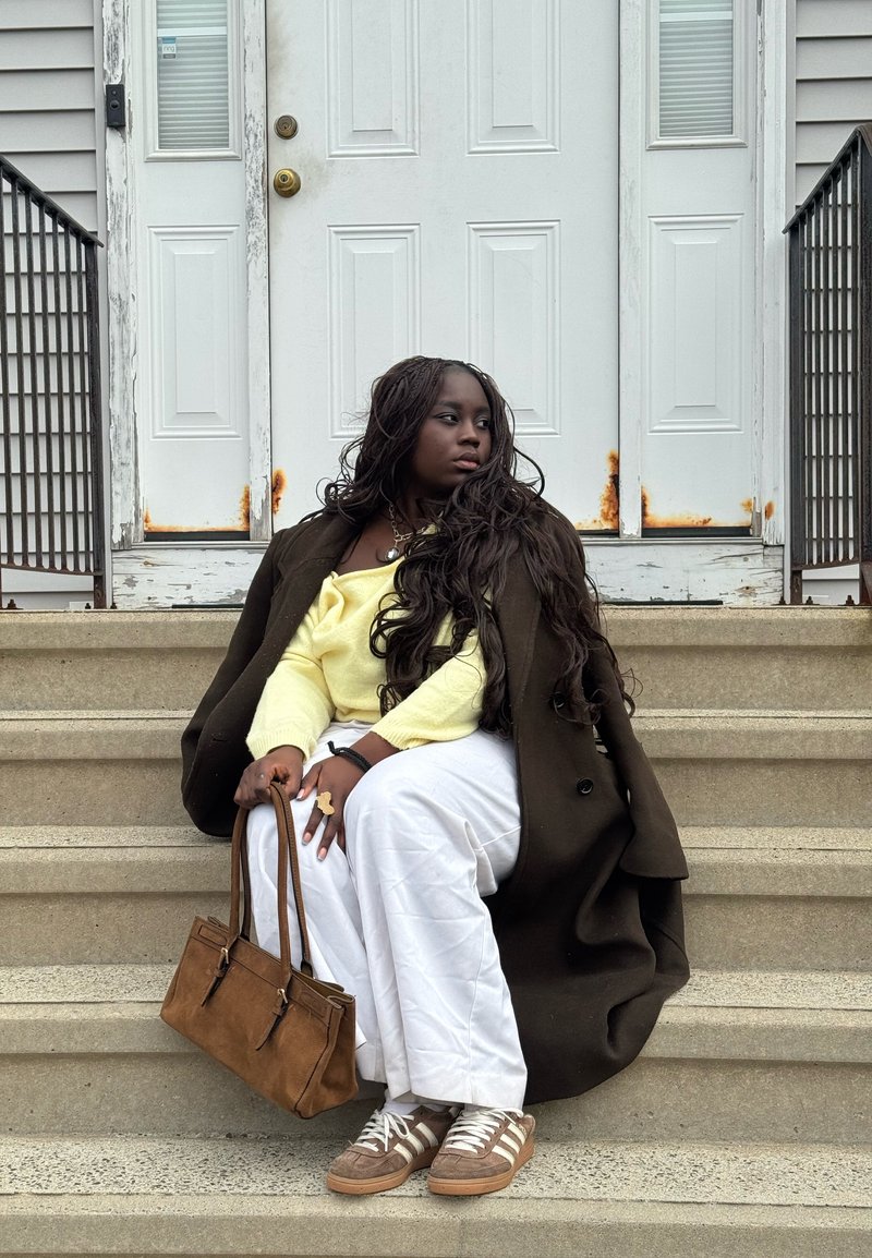 Personne assise en pantalon blanc, pull jaune, manteau foncé et baskets tenant un sac à main marron sur des marches avec des portes blanches rouillées derrière.