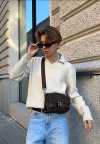 Καφέ δερμάτινη τσάντα ώμου με κούμπωμα, φορεμένη πάνω από ένα ανοιχτό κρεμ πουλόβερ; ανοιχτό μπλε τζιν παντελόνι; άτομο ρυθμίζει μαύρα γυαλιά ηλίου.