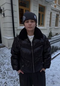 Doudoune noire à finition texturée avec col en fourrure. Portée sur une chemise blanche, associée à un pantalon sombre plissé et un bonnet gris.