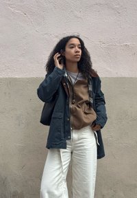 Jeune femme aux cheveux bouclés portant des vestes superposées et un pantalon blanc, debout devant un mur texturé bicolore beige et rose.