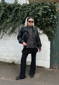 Čierna kožená bunda cez tmavý rebrovaný sveter, čierna sukňa s volánmi a rozšírené čierne nohavice. Doplnky zahŕňajú vzorovanú čelenku a slnečné okuliare.