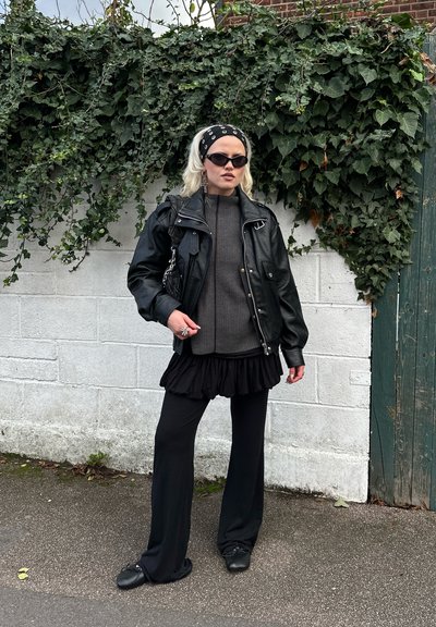 Chaqueta de cuero negra sobre un suéter oscuro de tejido acanalado, falda negra con volantes y pantalones negros acampanados. Accesorios: diadema estampada y gafas de sol.