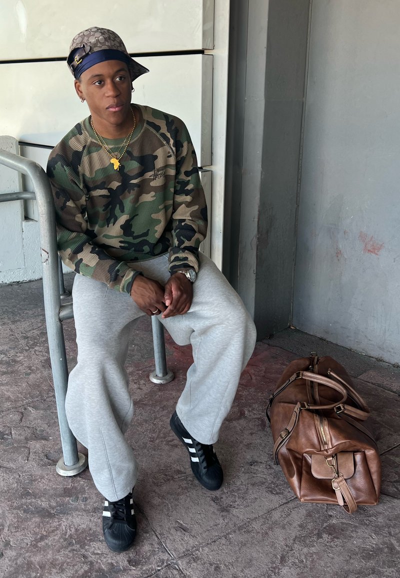Sweatshirt camuflado, calças de moletom cinzentas, ténis pretos com riscas brancas. Mala de viagem de couro castanho no chão ao lado de um indivíduo sentado.