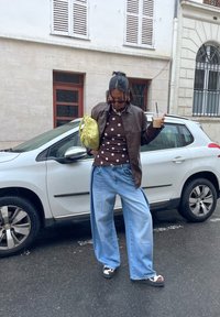 Veste en cuir marron, haut marron à pois, jean large bleu clair, sac à main jaune, chaussures noires et blanches ; scène de rue, voiture blanche garée.