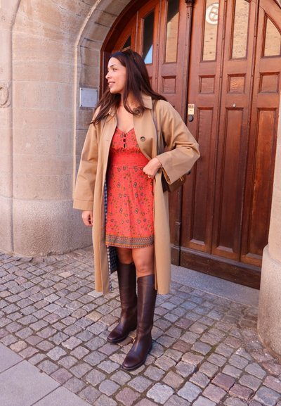 Mujer con abrigo beige, vestido rojo con estampado y botas marrón oscuro, de pie sobre adoquines frente a una puerta arqueada de madera.