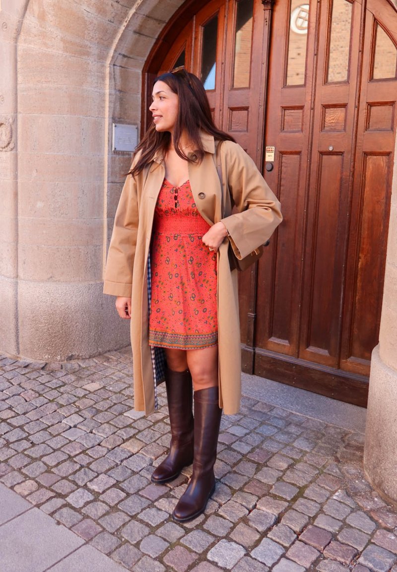 Femme portant un manteau beige, une robe rouge à motifs et des bottes marron foncé, debout sur un pavé devant une porte en bois arquée.