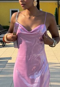 Abito in raso rosa con scollatura a drappo, spalline sottili e silhouette aderente. Accessoriato con una borsa a tracolla in argento e braccialetti.