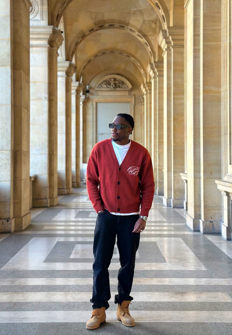 Homme portant un cardigan rouge, une chemise blanche, un pantalon noir et des bottes couleur beige se tient dans un corridor en pierre voûté avec un sol à motifs, regardant sur le côté.