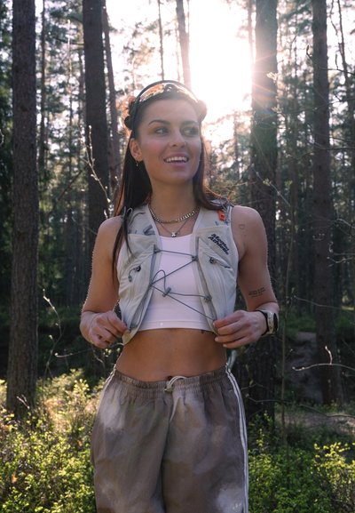 Mujer con crop top blanco y chaleco sonríe mientras ajusta las correas del chaleco en un bosque soleado con árboles altos y vegetación.