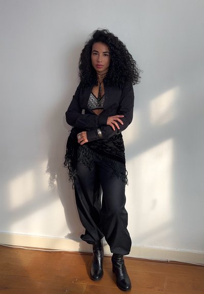 Blazer negro sobre un sujetador metálico, combinado con pantalones negros holgados y botas de tobillo. Una bufanda negra con flecos acentúa el conjunto.