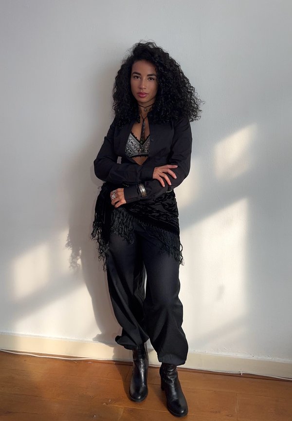 Blazer noir sur un bralette métallisé, associé à un pantalon noir ample et des bottines. Écharpe noire à franges souligne la tenue.