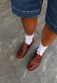 Ben med hvide ankelstrømper, brune læder snøresko og denimshorts der står på grå flisebelagt fortov.