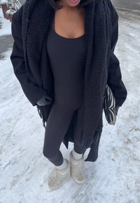 Personne vêtue d’une tenue entièrement noire avec manteau, écharpe et gants, tenant une pochette rayée zèbre, debout sur un sol enneigé.