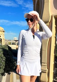 White zip-up athletic jacket paired with a matching skirt. Complemented by a black top with capped sleeves and sunglasses. Features a smooth texture and a fitted design.