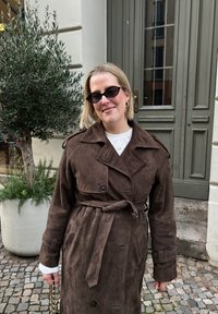 Femme portant un trench-coat marron foncé ceinturé et des lunettes de soleil noires, debout sur une rue pavée près d'un olivier en pot et d'une porte verte.