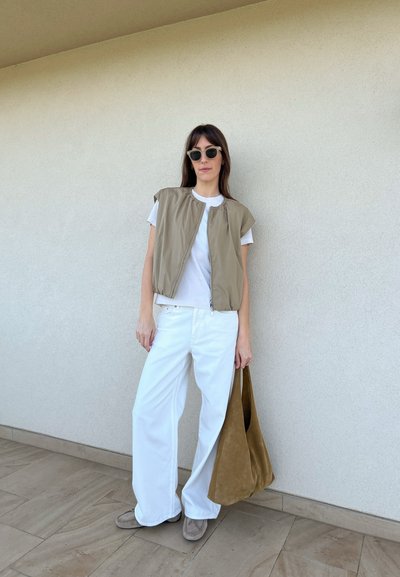 Chaleco ligero de color verde oliva, de mangas cortas, sobre una camiseta blanca, combinado con pantalones blancos de pierna ancha y un bolso de ante marrón. Unos zapatos grises completan el look.