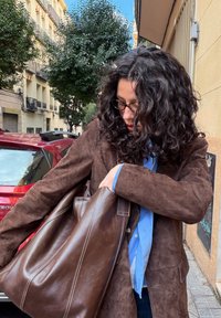 Femme aux cheveux bouclés et lunettes, portant un manteau marron, une chemise bleue, et tenant un grand sac en cuir marron sur un trottoir en ville.