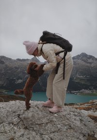 Žena v svetlom pufferovom kabáte a ružovej čiapke dvíha hnedého pudla proti skalnatej krajine s horami a jazerom v pozadí.