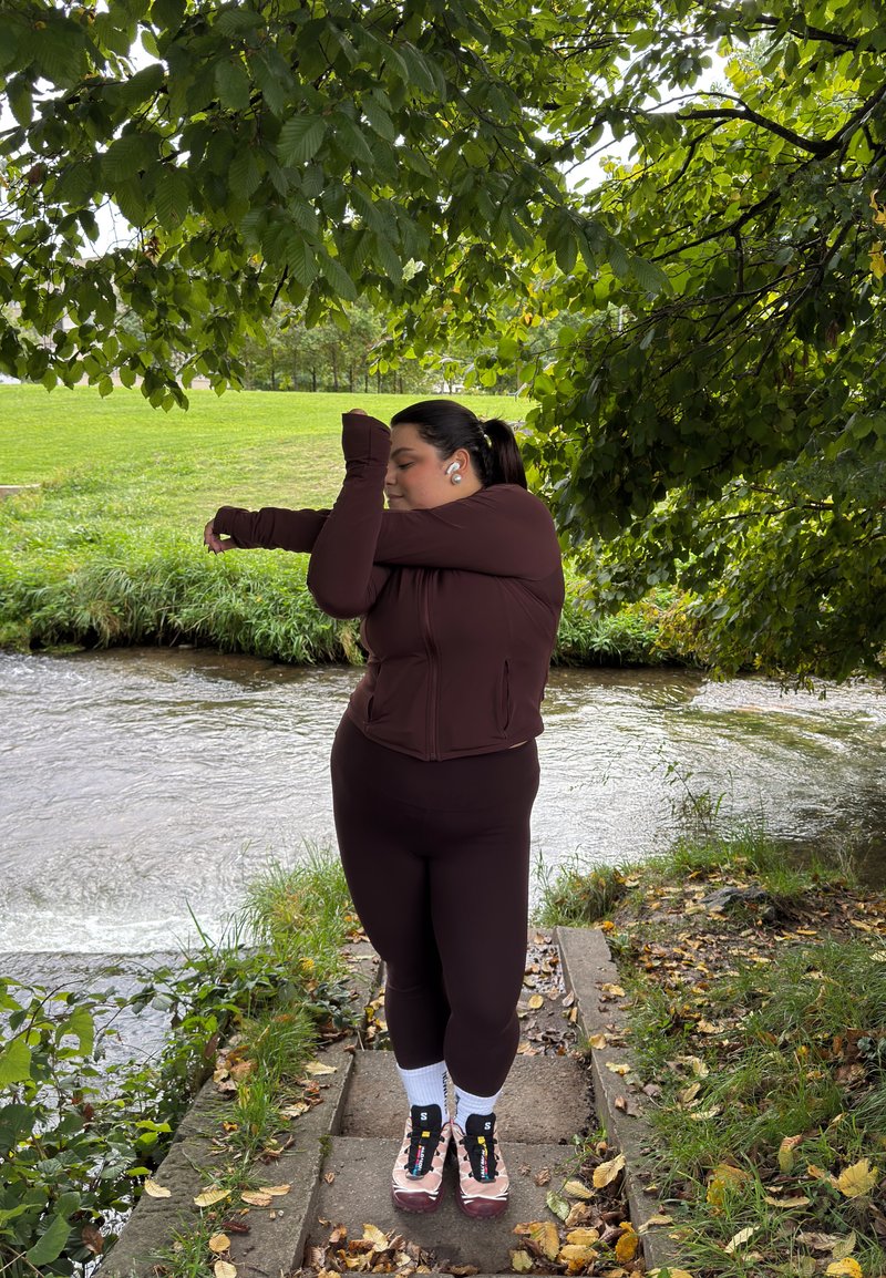 Mulher em roupa de desporto bordô esticando o braço junto a um rio sob ramos de árvores, num caminho gramado com folhas caídas.