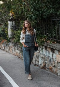 Salopette en denim grise avec une coupe décontractée, associée à une chemise blanche. Chaussures beige à enfiler. Mur en pierre naturelle et verdure en arrière-plan.