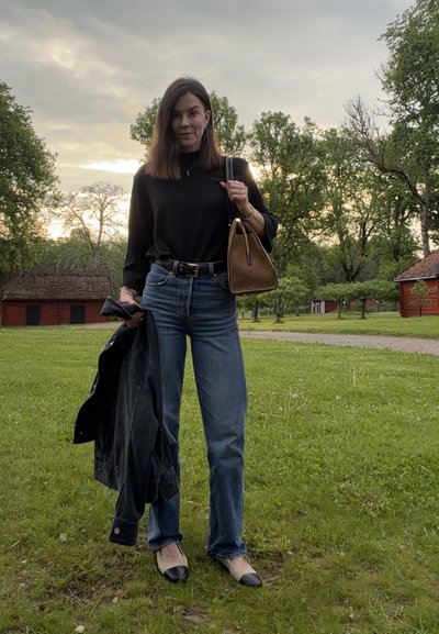 Mujer de pie sobre la hierba sosteniendo una chaqueta en una mano y un bolso marrón en el hombro, con árboles y edificios rojos al fondo.