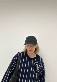 Yli suuri tummansininen ja valkoinen raidallinen neuletakki, jossa on iso 'G' -patch, yhdistetty mustaan lippikseen. Henkilöllä on olkalaukku.