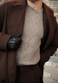 Homme portant un manteau marron, un pull beige texturé, un pantalon marron et un gant en cuir noir faisant un pouce levé près d'un mur en briques.
