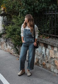 Salopette en denim bleu clair, avec des jambes larges et des ourlets retroussés. Associée à un blouse crème et des chaussures à enfiler beiges, sur un fond de mur en pierre.