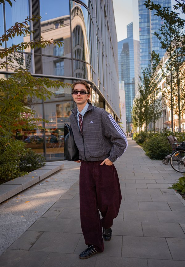 Un hombre con gafas de sol camina por una acera de la ciudad y lleva una bolsa negra entre edificios modernos y árboles.