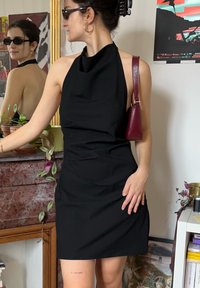Femme aux cheveux foncés attachés en chignon, portant une robe noire dos nu, des lunettes de soleil et un sac à main rouge, debout à l'intérieur près d'un miroir et de plantes.