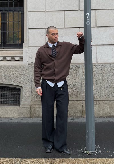 Cárdigan marrón sobre una camisa blanca y corbata negra, combinado con pantalones negros sueltos. De pie cerca de un poste de luz gris, con un fondo de concreto.