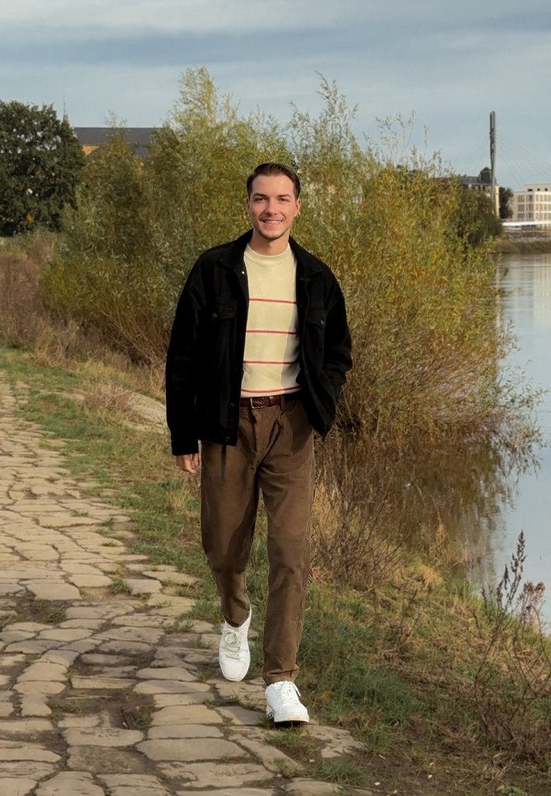 Черно яке върху светъл кремав пуловер с червени ивици, кафяви панталони и бели маратонки, крачещи по каменна пътека до вода и зеленина.