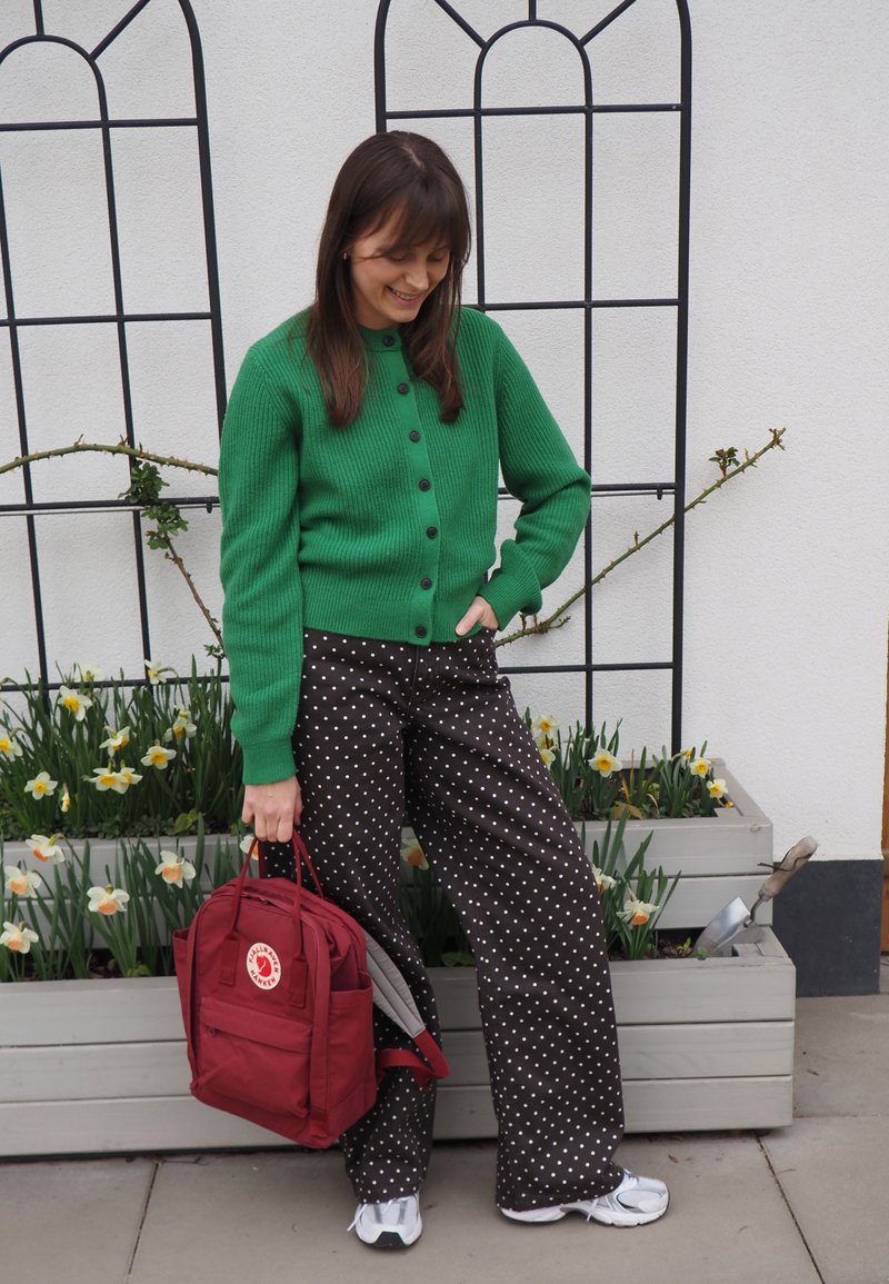 Femme en cardigan vert et pantalon noir à pois, tenant un sac à dos rouge, debout près de jardinières avec des jonquilles contre un mur blanc.