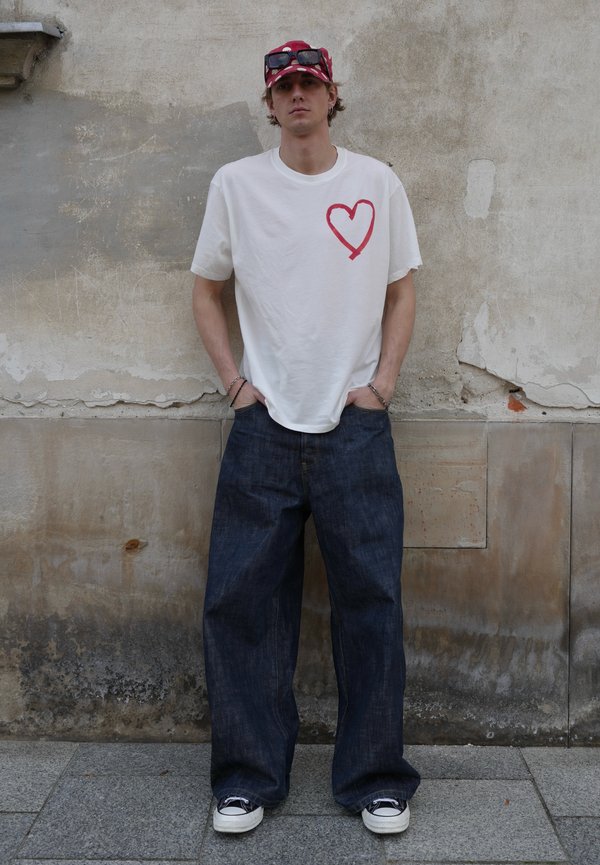 Joven con camiseta blanca con diseño de corazón rojo, vaqueros anchos oscuros, gorra roja con gafas de sol, de pie junto a una pared de hormigón desgastada.
