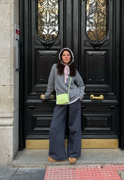Suéter gris con cordones rosas, pantalones anchos a rayas y un bolso bandolera verde. De pie frente a una puerta negra ornamentada.