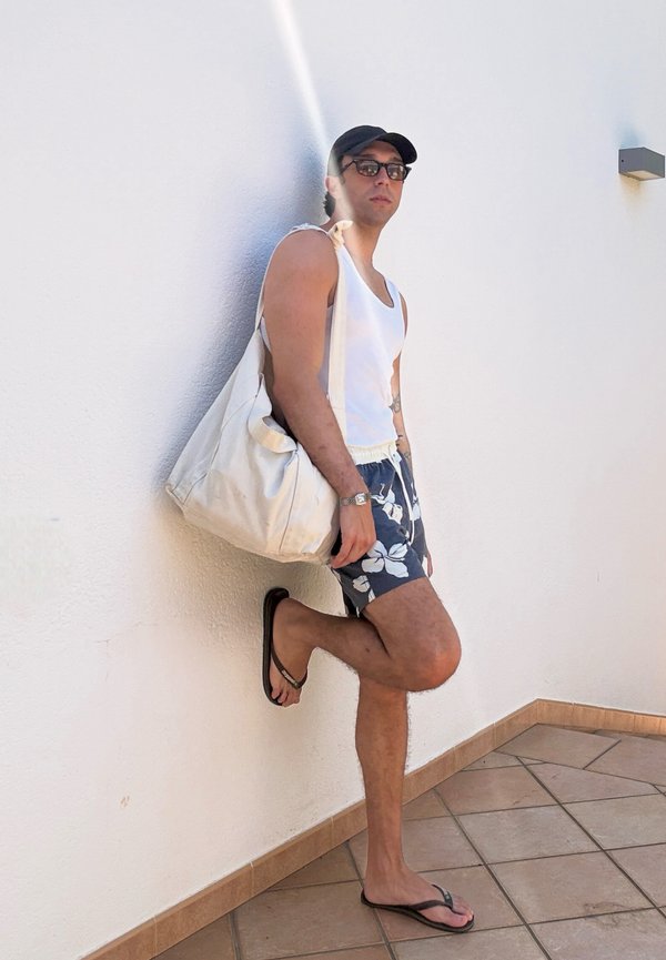 Mann in weissem Tanktop, geblümten Shorts, schwarzer Kappe und Sonnenbrille lehnt an einer weissen Wand und hält eine grosse beige Tasche.